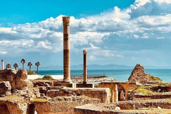 De hautes colonnes de pierre antiques et des ruines se dressent sur une côte, surplombant la mer sous un ciel bleu vif et nuageux.
