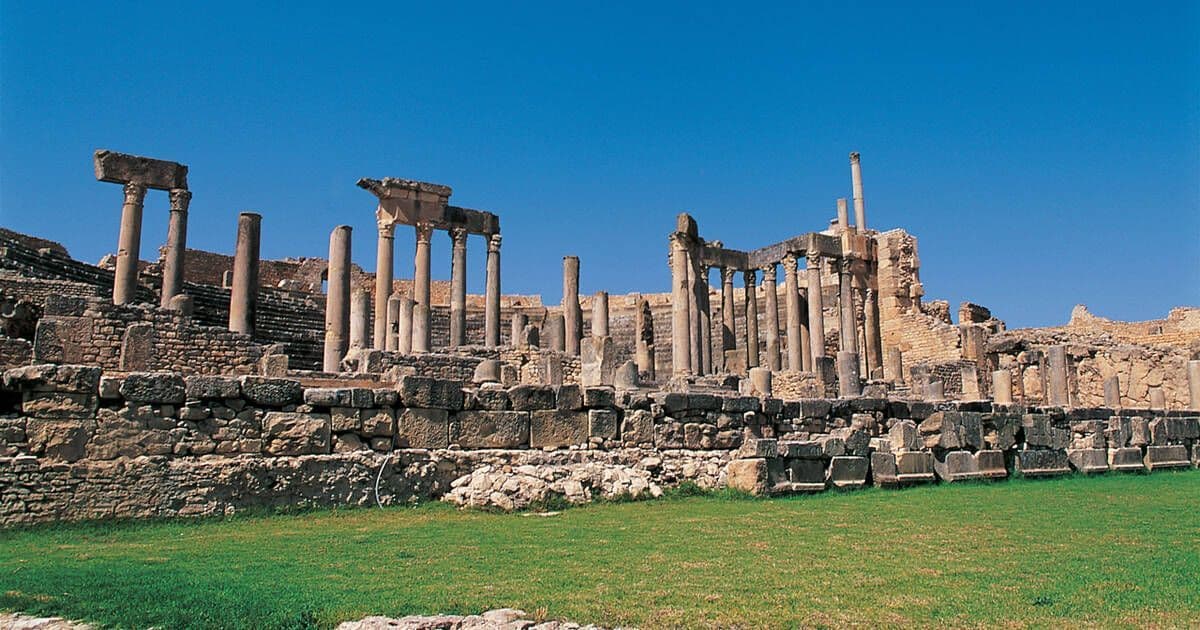 D'anciennes ruines de pierre avec de nombreuses colonnes et des murs se dressent sur une colline surplombant une pelouse verte sous un ciel bleu clair.