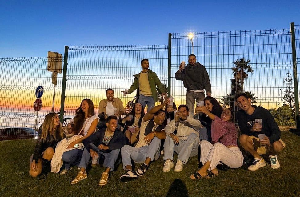 Un gruppo WeRoad in viaggio sorride e posa per una foto su un pendio erboso al tramonto, tenendo in mano delle bevande davanti a una recinzione.