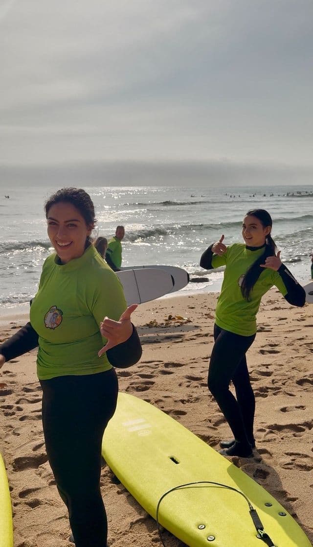 Due donne di un viaggio di gruppo WeRoad, in muta, sorridono e fanno il segno shaka in spiaggia con le loro tavole da surf.