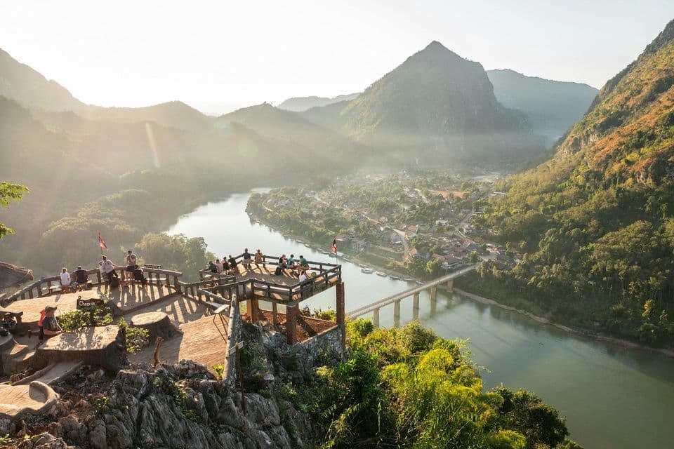 Un gruppo WeRoad in viaggio su un belvedere in legno, con vista su un fiume serpeggiante, un villaggio e montagne velate all'alba.