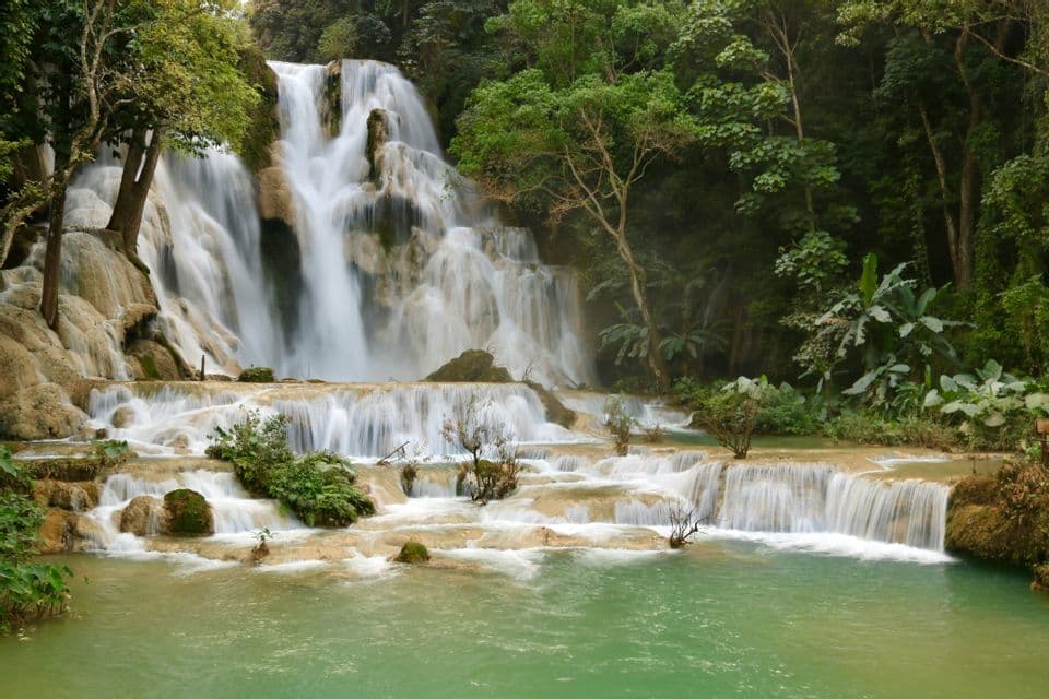 Una cascata a più livelli si getta su rocce chiare in una piscina naturale turchese, avvolta da una lussureggiante foresta verde.