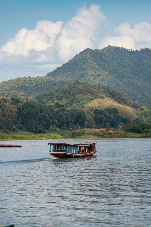 Una barca di legno blu e marrone attraversa un fiume con una grande montagna coperta di foreste sullo sfondo sotto un cielo parzialmente nuvoloso.
