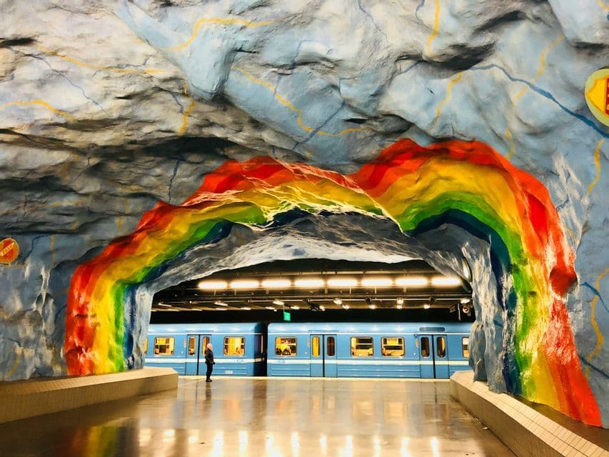 Un treno blu è fermo in una stazione con pareti simili a caverne, dove un grande arcobaleno è dipinto sopra un arco.