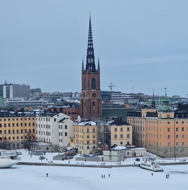 Un campanile storico si affaccia su una città coperta di neve e un fiume ghiacciato dove le persone camminano.