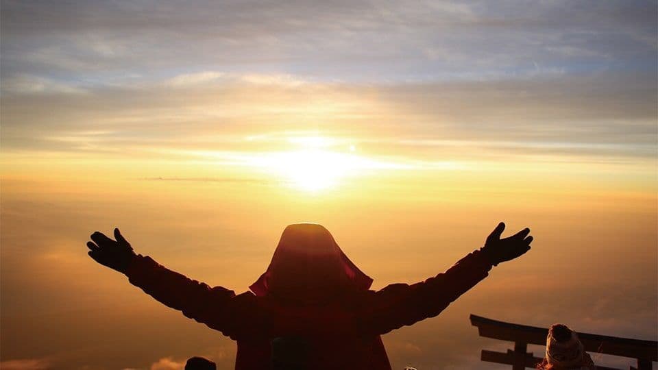 Una persona con una giacca rossa con cappuccio, vista da dietro, alza le braccia verso l'alba su un mare di nuvole.