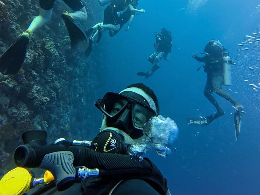 Una persona in muta subacquea si scatta un selfie sott'acqua durante un viaggio di gruppo WeRoad, con altri subacquei che nuotano vicino a una barriera corallina.