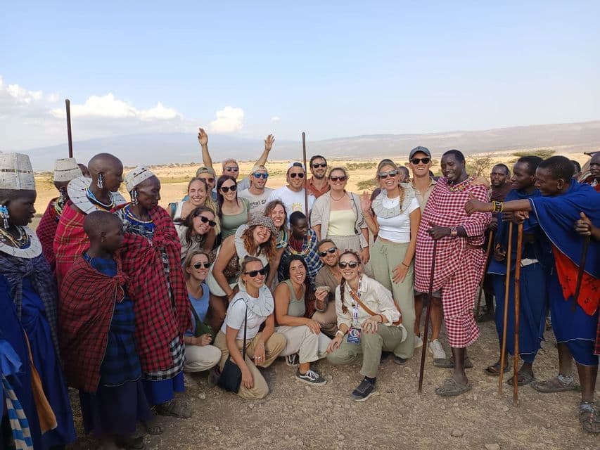 A WeRoad group trip poses for a photo with members of a local community dressed in traditional attire in a savanna.