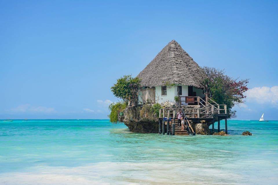 A building with a thatched roof sits on a rock in the turquoise ocean under a clear blue sky.