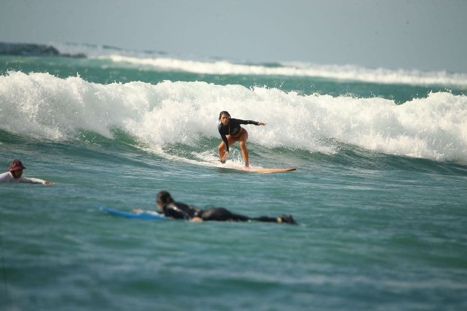 Una donna con una muta nera cavalca un'onda sulla sua tavola da surf, con altri due surfisti in acqua nelle vicinanze.