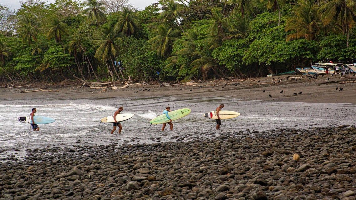 Quattro surfisti di un gruppo WeRoad, con le loro tavole, escono dall'acqua su una spiaggia rocciosa con una fitta giungla sullo sfondo.