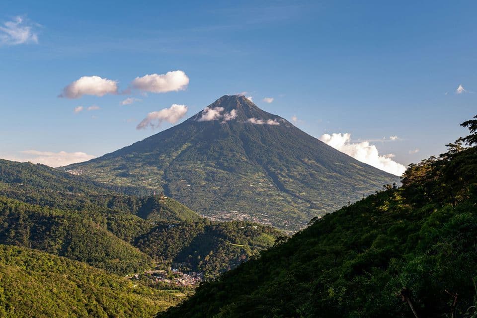 Un vulcano verde conico con nuvole attorno alla sua cima, che domina una valle con una piccola città e fitte foreste sotto un cielo azzurro.
