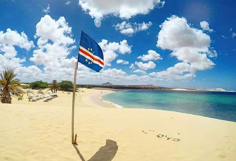 La bandiera di Capo Verde è piantata su una duna di sabbia, con vista su una spiaggia tropicale con acqua turchese sotto un cielo parzialmente nuvoloso.