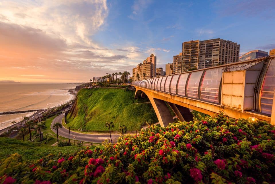Un ponte pedonale coperto su una scogliera verde che si affaccia su una città costiera e sull'oceano durante un tramonto colorato.