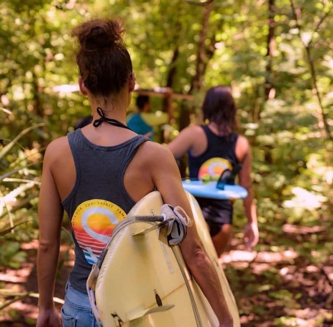A WeRoad group trip of surfers carries their boards while walking on a path through a sunlit green forest.