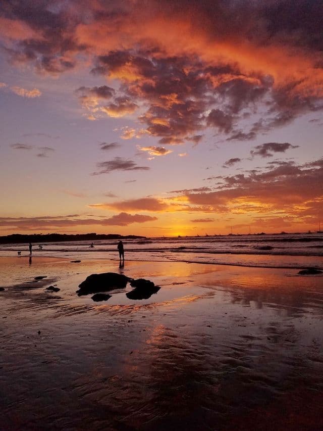 Silhouetten von Menschen an einem nassen Strand bei Ebbe, mit einem farbenfrohen orange-lila Sonnenuntergang, der sich in Sand und Wasser spiegelt.