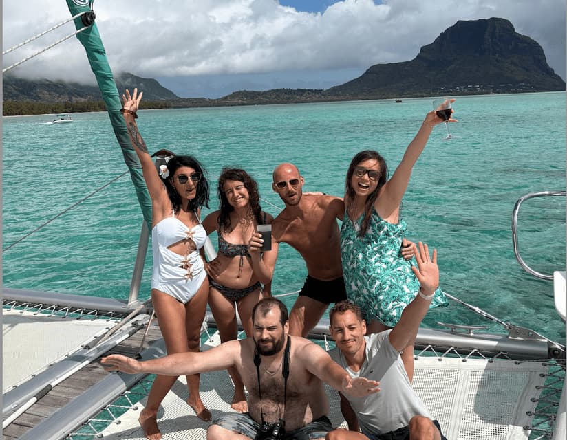 Un groupe WeRoad en voyage posant joyeusement sur le filet d'un catamaran, naviguant sur des eaux turquoises avec une île montagneuse en arrière-plan.