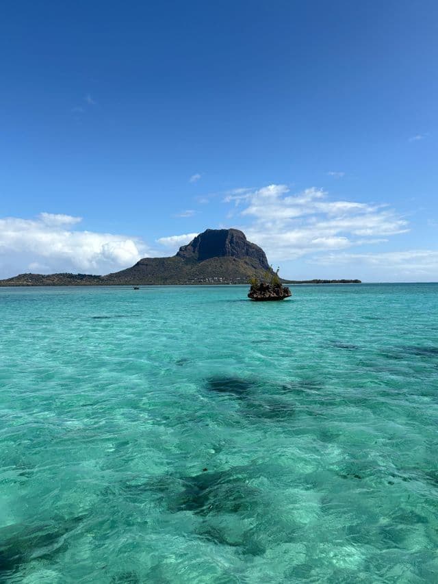 Une grande montagne se dresse au loin au-delà d'une mer claire et turquoise, avec un petit îlot rocheux au premier plan sous un ciel bleu.