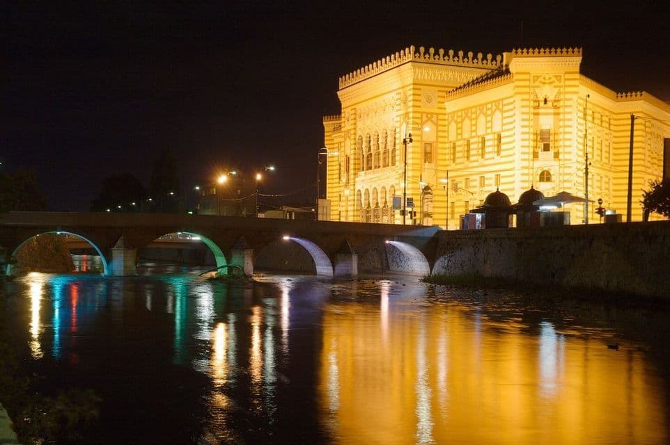 Un edificio giallo sontuoso e un ponte di pietra sono illuminati di notte, con le loro luci riflesse sul fiume.