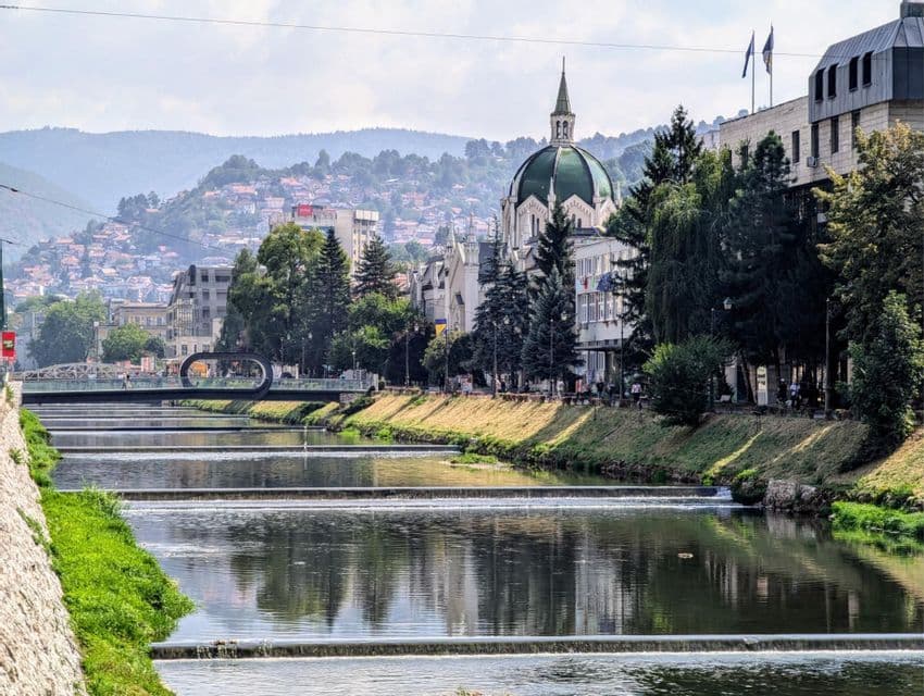 Un fiume scorre su piccoli sbarramenti attraverso una città con un edificio dalla cupola verde e una collina ricoperta di case.