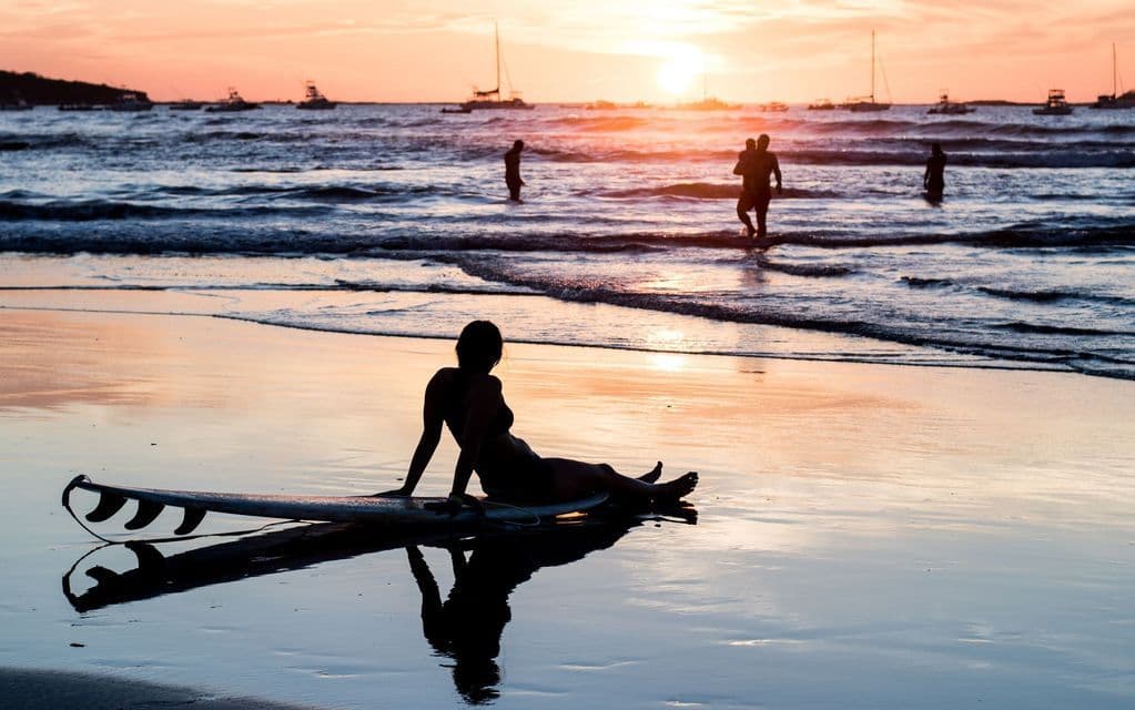Eine Frau sitzt als Silhouette auf einem Surfbrett im feuchten Sand eines Strandes bei Sonnenuntergang, mit Booten am Horizont.
