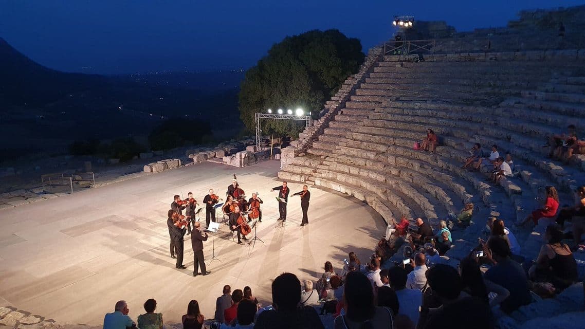 Una orquesta de cuerda actúa en el escenario para una audiencia sentada en un gran anfiteatro de piedra al atardecer.