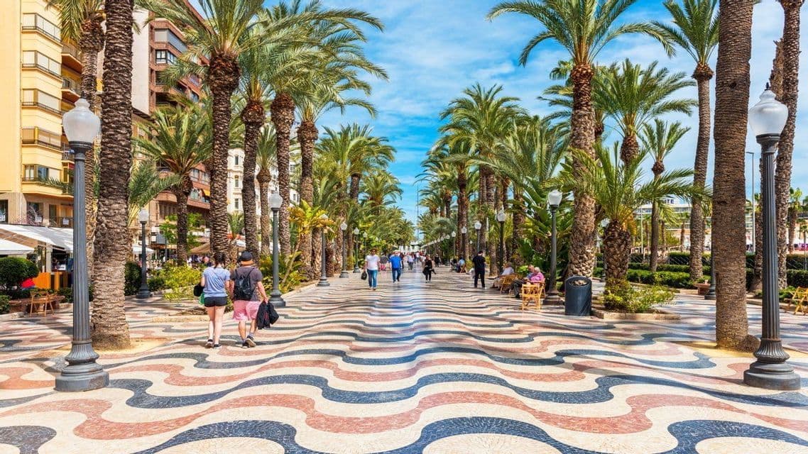 Le persone passeggiano lungo un ampio lungomare pavimentato con un motivo a mosaico ondulato e colorato e fiancheggiato da alte palme in una giornata di sole.