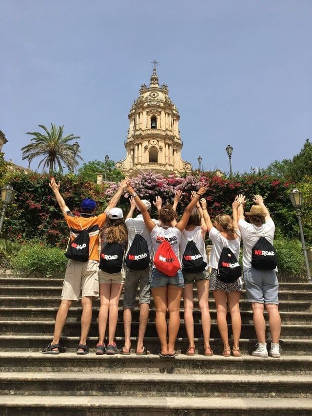 A WeRoad group trip seen from behind, standing on stone steps with arms raised towards an ornate church with a bell tower.