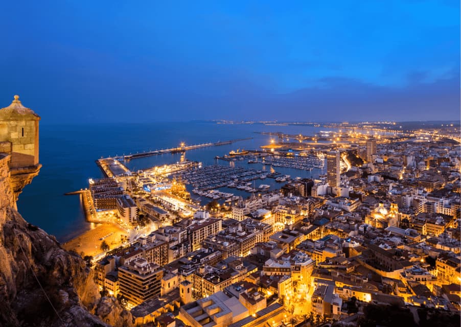 Una vista sopraelevata di una città costiera al tramonto, con i suoi edifici illuminati, il porto e una torretta in pietra in primo piano.