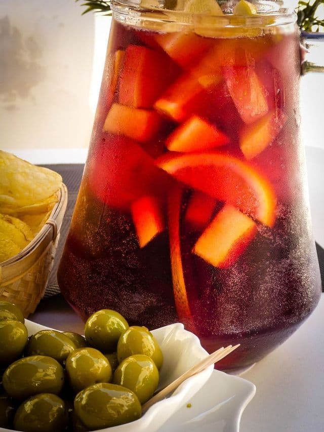 Un primo piano di una brocca di sangria rossa con frutta fresca accanto a una ciotola di olive verdi e un cestino di patatine su un tavolo.