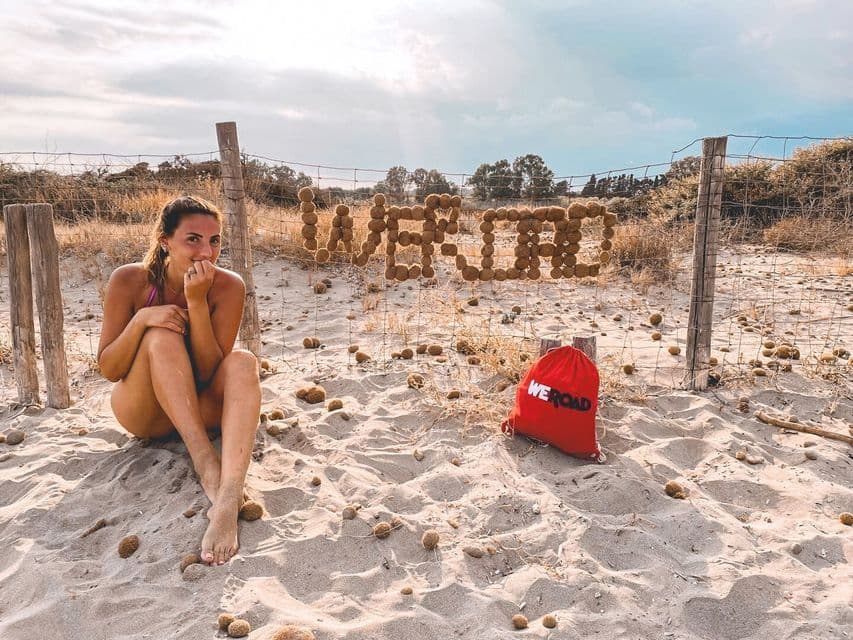 Una mujer en bikini descansa en la arena junto a una valla de alambre con el logo de WeRoad, hecho de bolas de algas marinas, y una bolsa roja de la marca.
