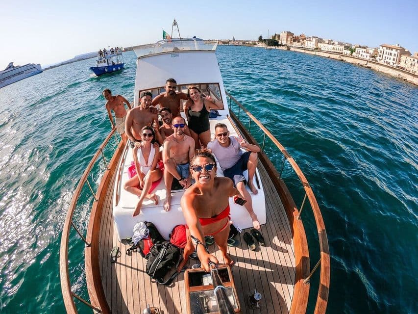Un grupo de WeRoad de viaje sonríe para una selfie en la cubierta de madera de un barco, navegando por aguas azules cerca de una ciudad costera.