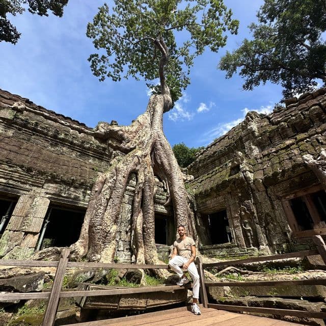 A low-angle shot of a man sitting on a wooden railing in front of ancient temple ruins being overtaken by the roots of a large tree.