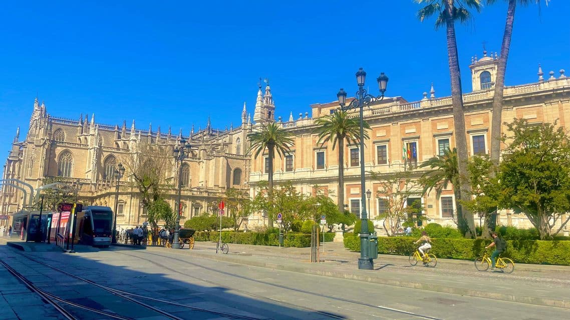 Una grande cattedrale riccamente decorata e un edificio storico fiancheggiano una via cittadina soleggiata, con un tram, palme e ciclisti.