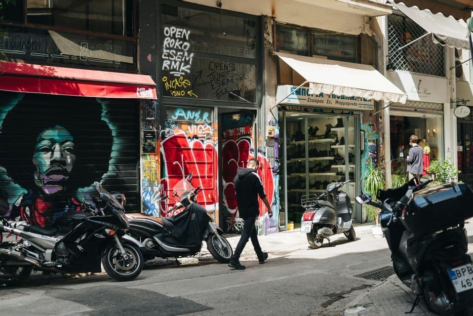 Una persona con una sudadera con capucha negra camina por una calle de la ciudad bordeada de escaparates cubiertos de grafitis y motocicletas estacionadas.
