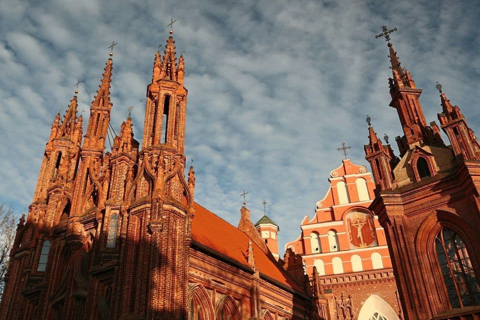 Una vista dal basso di due intricate chiese gotiche in mattoni rossi con alte guglie sotto un cielo blu nuvoloso.