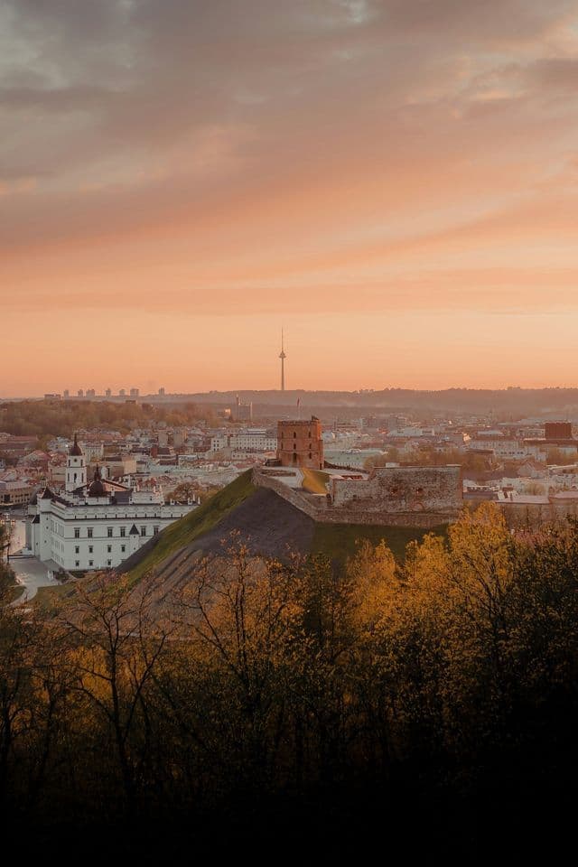 Una torre di mattoni su una collina verde domina un paesaggio urbano con una torre TV in lontananza sotto un caldo cielo al tramonto.