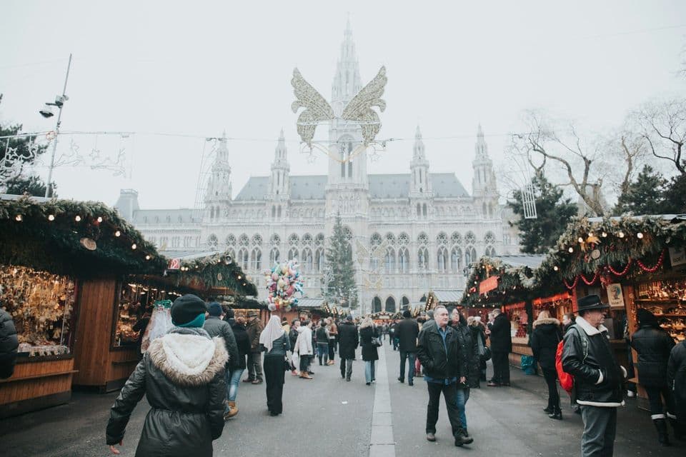 Una folla di persone passeggia in un mercatino di Natale all'aperto con bancarelle di legno di fronte a un grande edificio ornato.