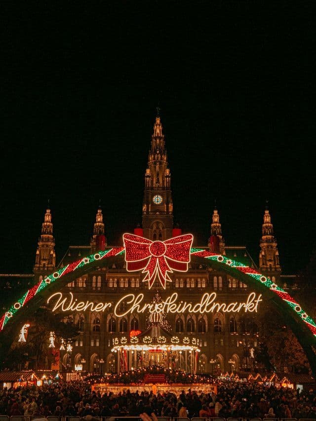 L'arco d'ingresso illuminato del 'Wiener Christkindlmarkt' davanti a un grande edificio, con folle di persone che si godono il mercato di notte.