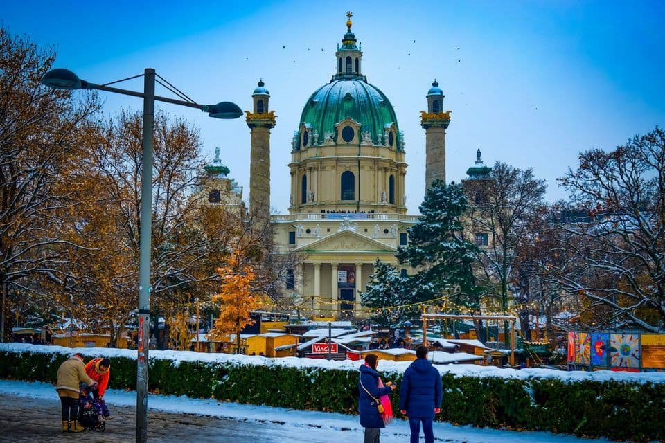 Una grande chiesa con una cupola verde domina un parco innevato dove le persone passeggiano per un mercatino invernale.