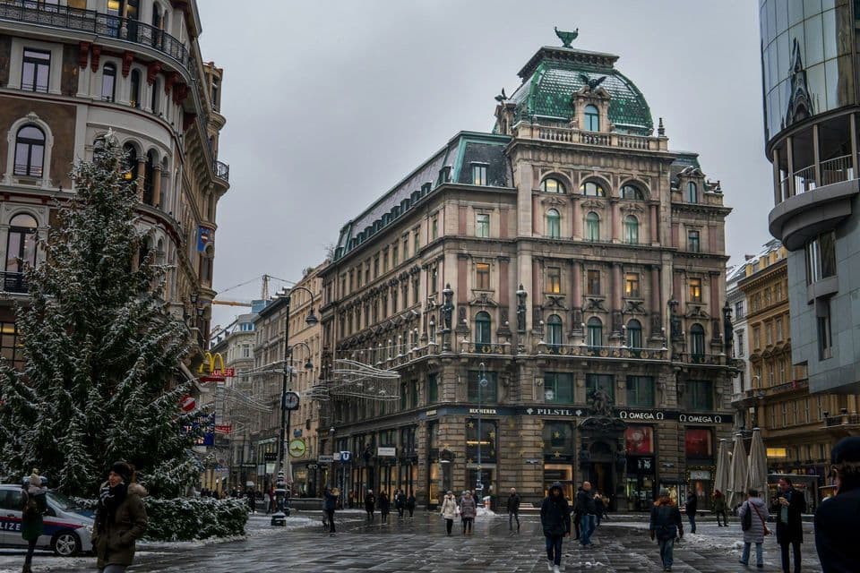 Fußgänger gehen über einen verschneiten Stadtplatz mit einem großen Weihnachtsbaum und prächtigen historischen Gebäuden unter bewölktem Himmel.