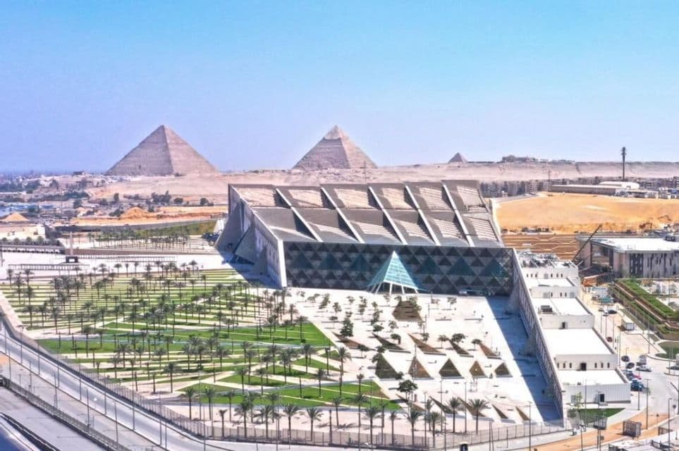 Un museo moderno con giardini paesaggistici e palme, con tre antiche piramidi visibili sullo sfondo del deserto sotto un cielo azzurro e limpido.