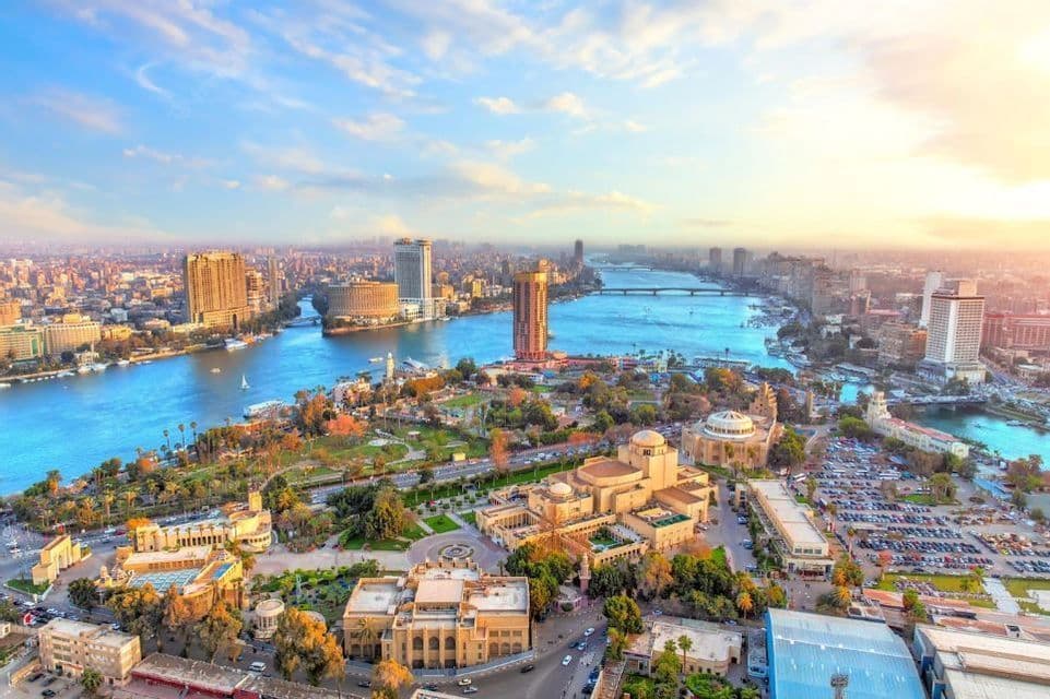 Una vista aerea di una vasta città tentacolare attraversata da un ampio fiume blu, fiancheggiata da edifici moderni e parchi verdi sotto un cielo soleggiato.