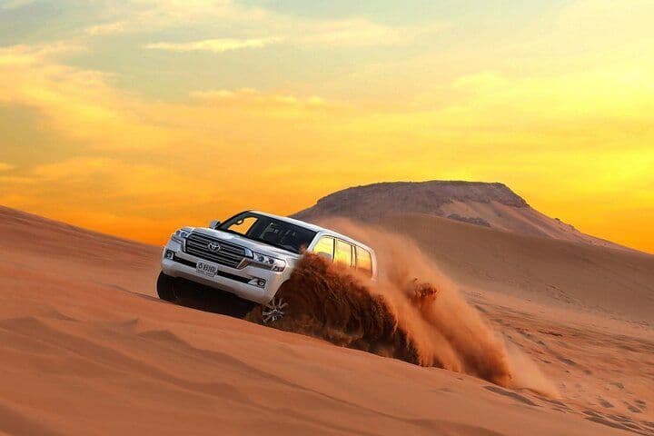 Un SUV bianco scende una grande duna di sabbia nel deserto, sollevando una nuvola di sabbia contro un cielo dorato al tramonto.