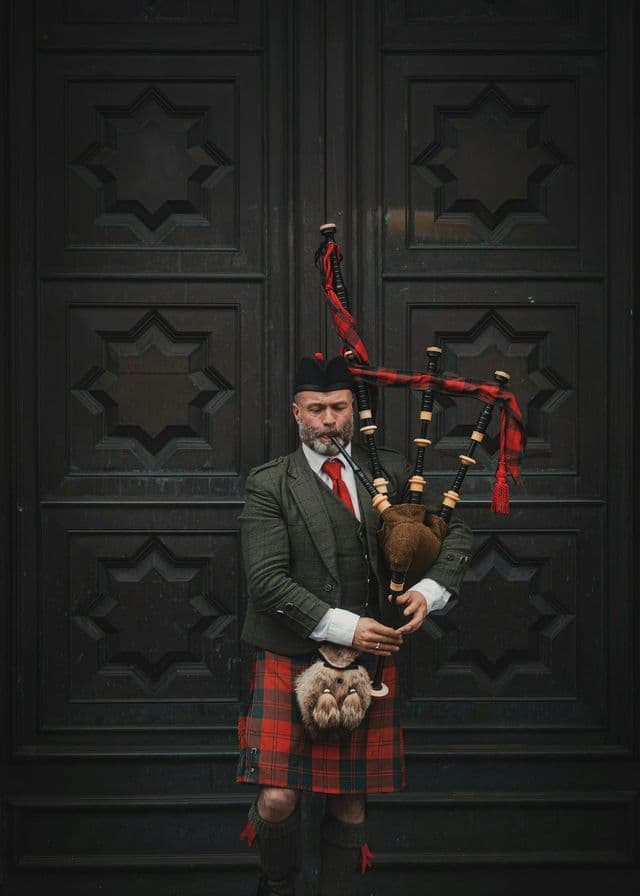 Ein Mann in traditioneller schottischer Tracht, darunter ein roter Tartan-Kilt, hält einen Dudelsack, während er vor einer verzierten dunklen Tür steht.