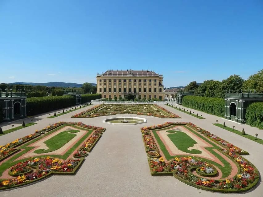 Un gran palacio amarillo al final de un vasto jardín formal con intrincados y coloridos parterres y caminos bajo un cielo azul claro.