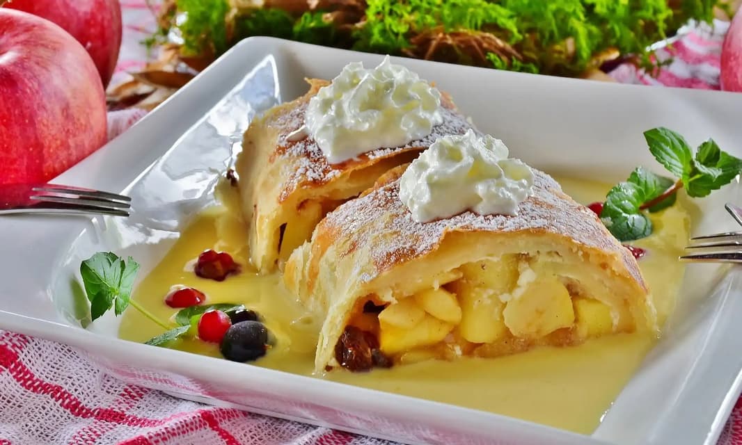Dos rebanadas de strudel de manzana en un plato blanco, coronadas con crema batida y azúcar glas, servidas sobre un lecho de salsa de vainilla.
