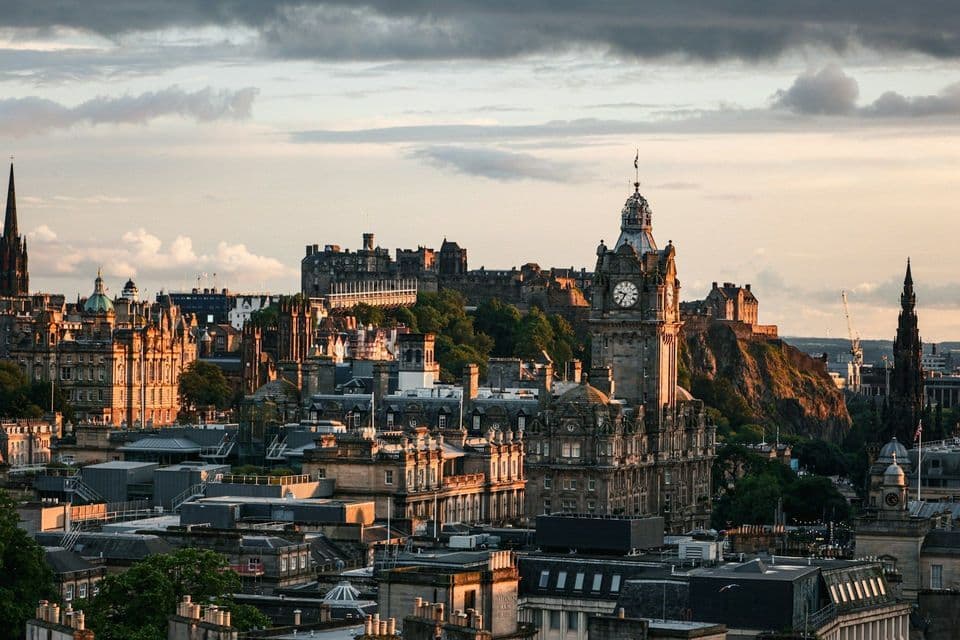 A historic city skyline featuring a prominent clock tower and a castle on a hill, bathed in the warm light of sunset.