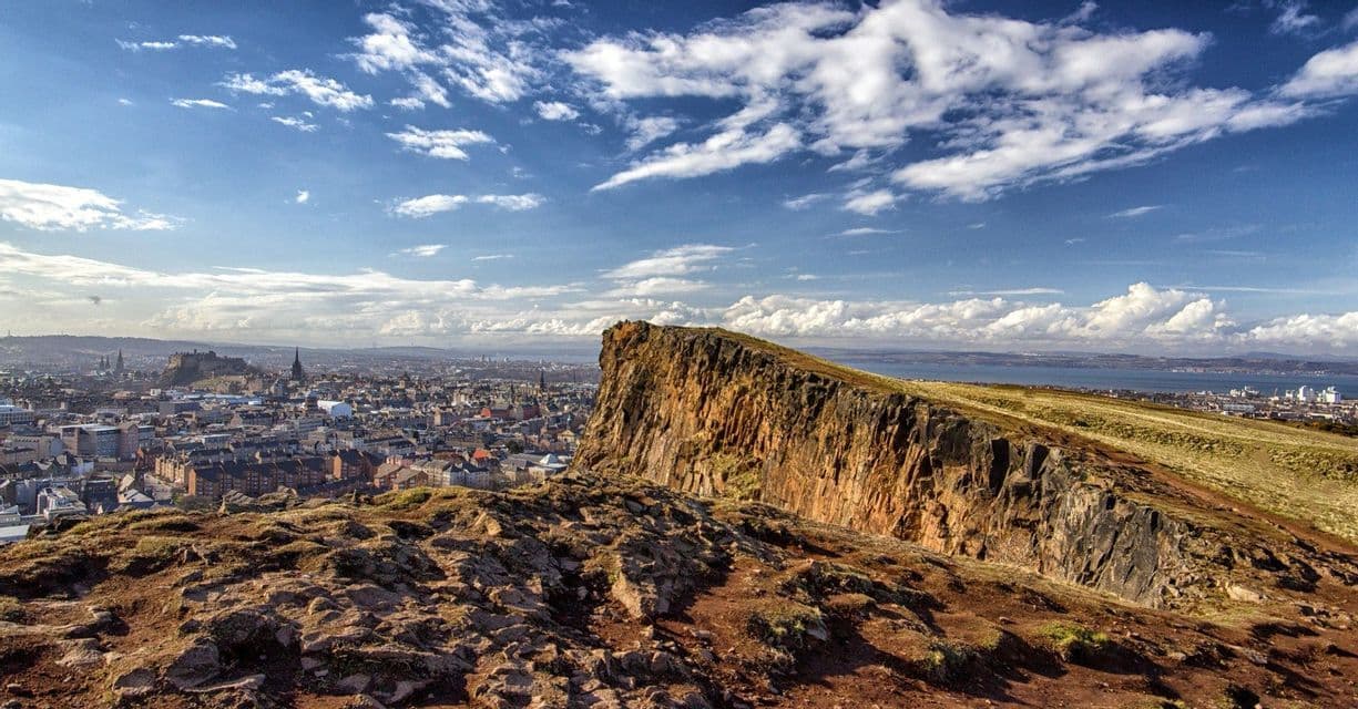 A panoramic view from a rocky cliff overlooking a city and a bay under a blue sky with white clouds.