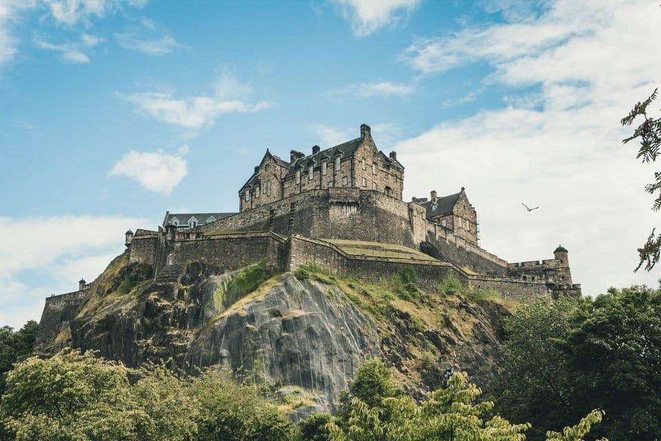 A large stone castle sits atop a steep, rocky cliff, framed by green trees below and a partly cloudy blue sky above.